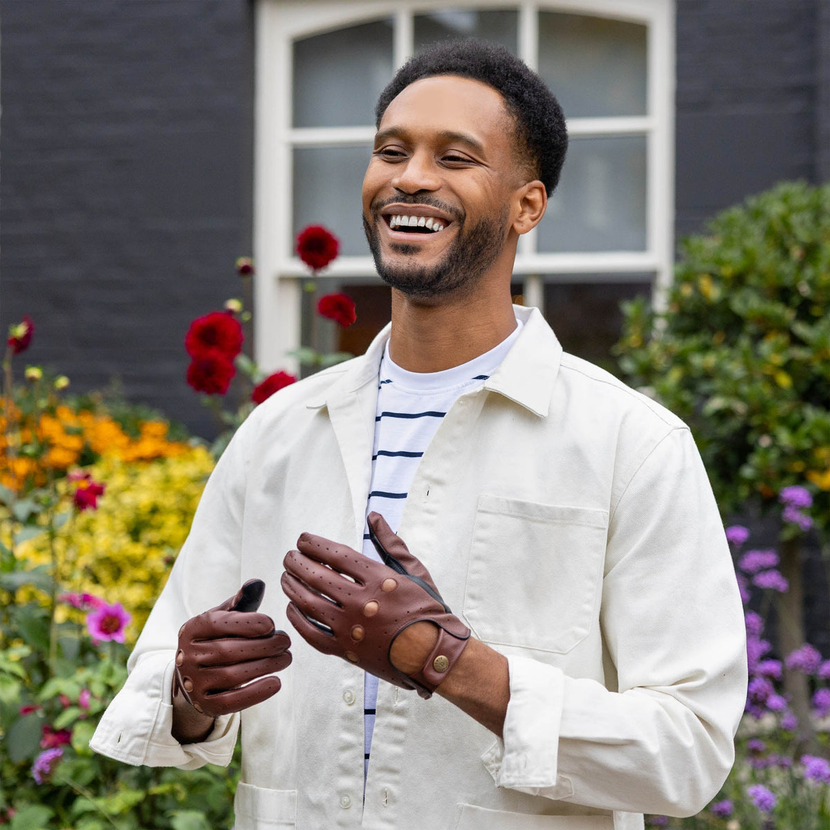 Man wearing a white jacket and leather driving gloves in english tan/black standing in front of a building with flowers.